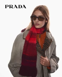 Model wearing dark sunglasses, an orange sweater, a grey coat, and a red knitted scarf against a white background