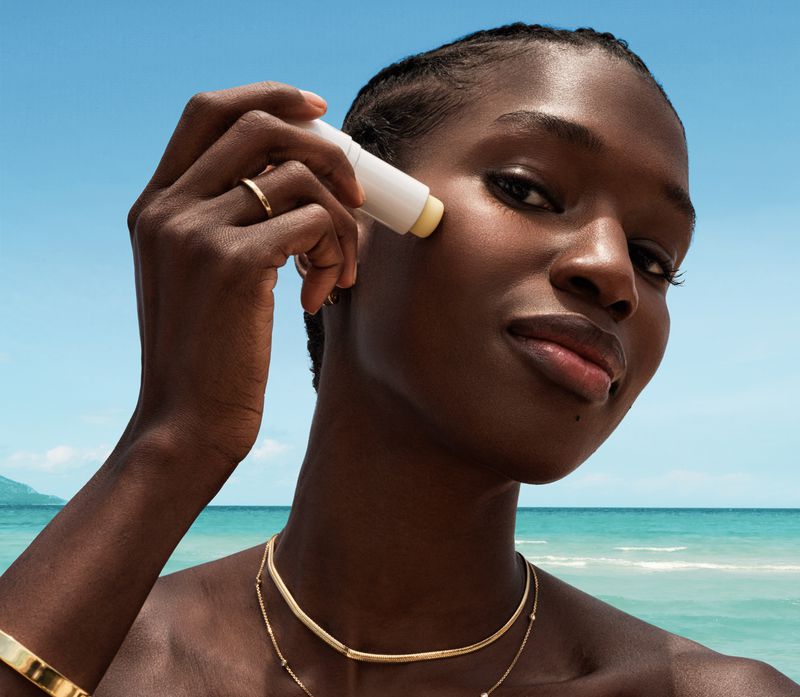 An African-descent woman on a beach applies a skincare stick to her cheek, with a blue sky and ocean in the background on a sunny day.