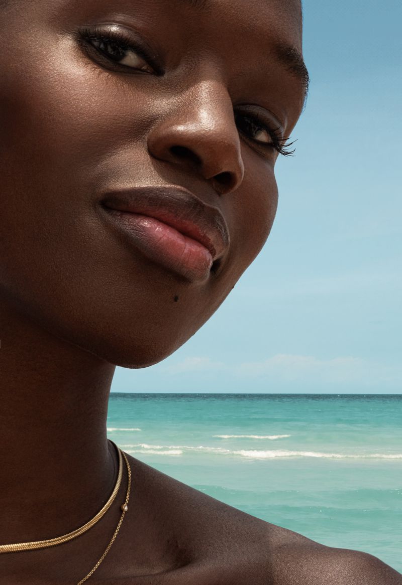 Close-up portrait of a Black woman with golden jewelry in front of a turquoise ocean and clear blue sky, capturing beauty and serenity.