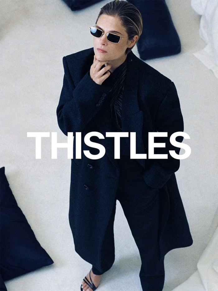 Louisa Jacobson in a black coat and sunglasses posing for the THISTLES AW26 campaign on a white floor