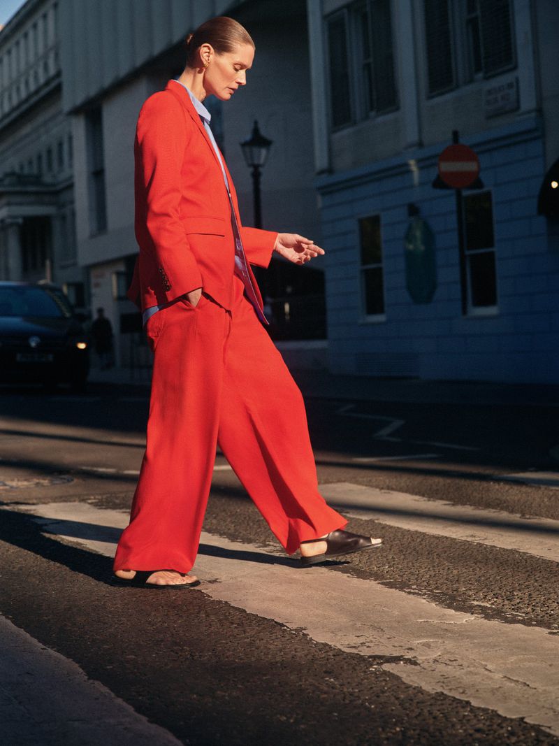 Adult woman in a red pantsuit strides across a crosswalk in an urban setting on a sunny day, showcasing a blend of fashion and city life.