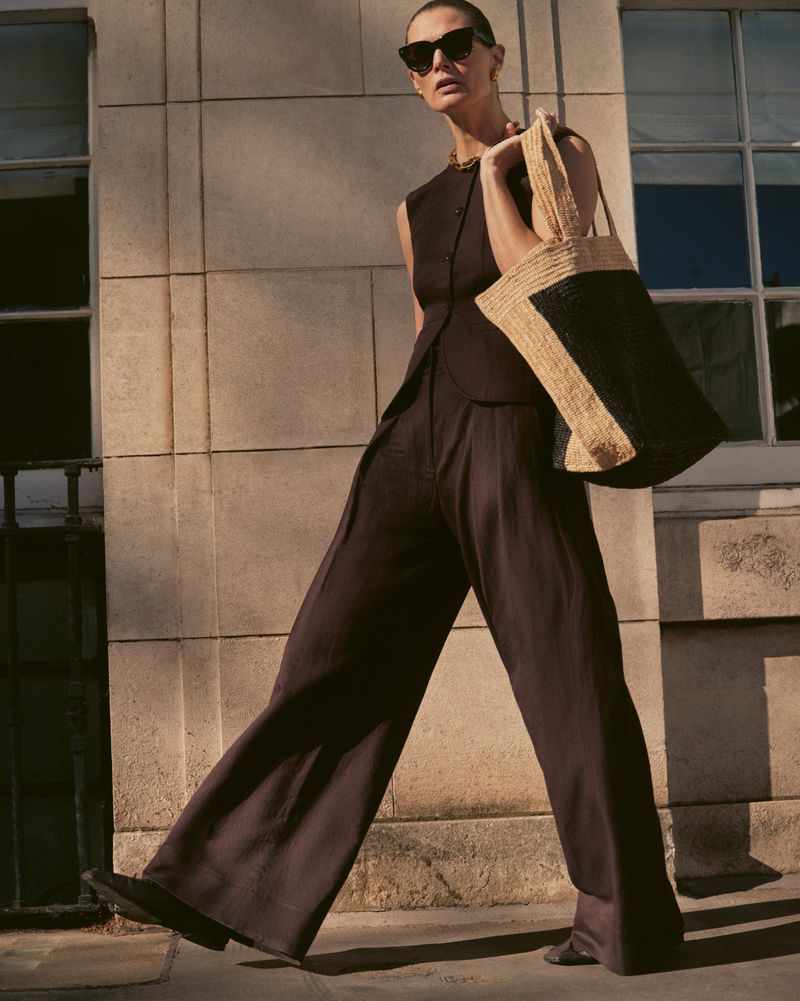 A full shot of a model walking outside wearing a sleeveless top, wide-leg trousers, sunglasses, and carrying a large tote bag.