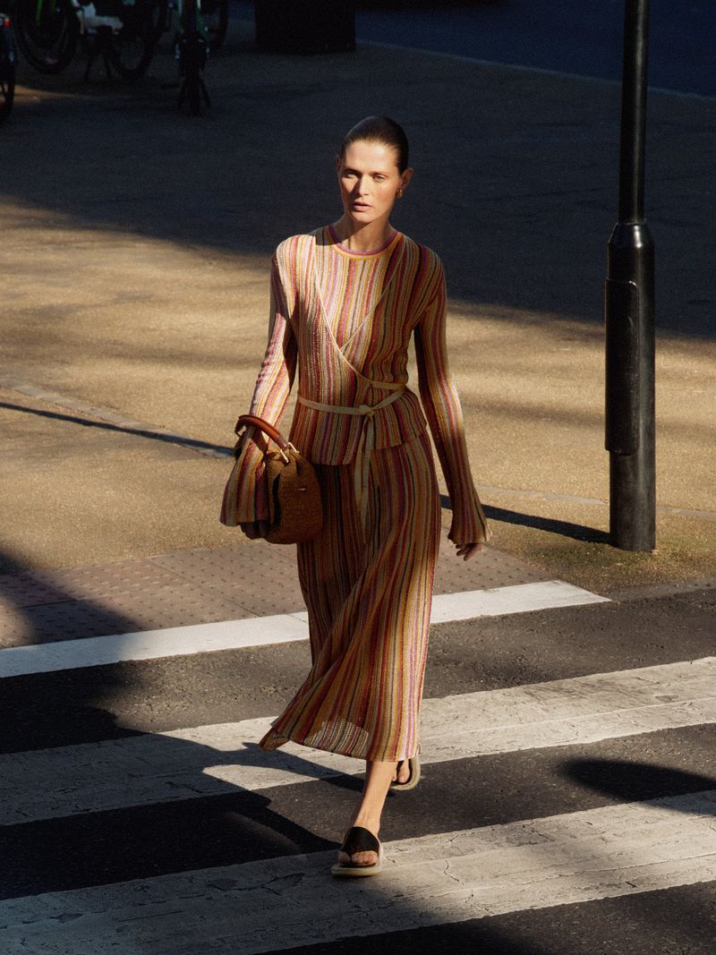 A model walks across a crosswalk in a striped midi dress and carrying a handbag on a city street with diffused natural lighting.
