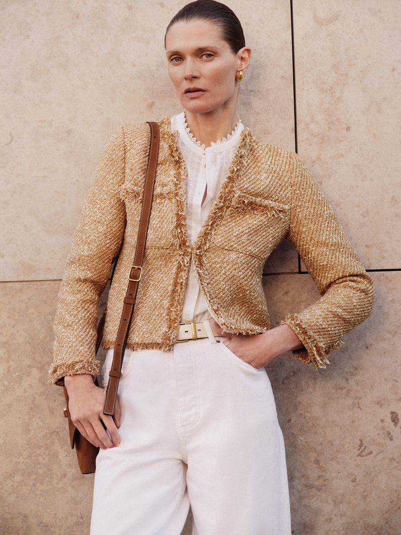 Woman in a tweed jacket and white pants stands against a textured wall with a brown crossbody bag. Fashion, clothing, style, and elegance.