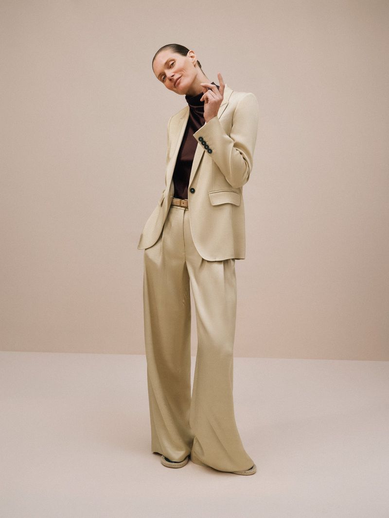 A slender woman poses in a cream-colored blazer and wide-leg pants, a brown top peeking beneath the blazer, against a neutral background.
