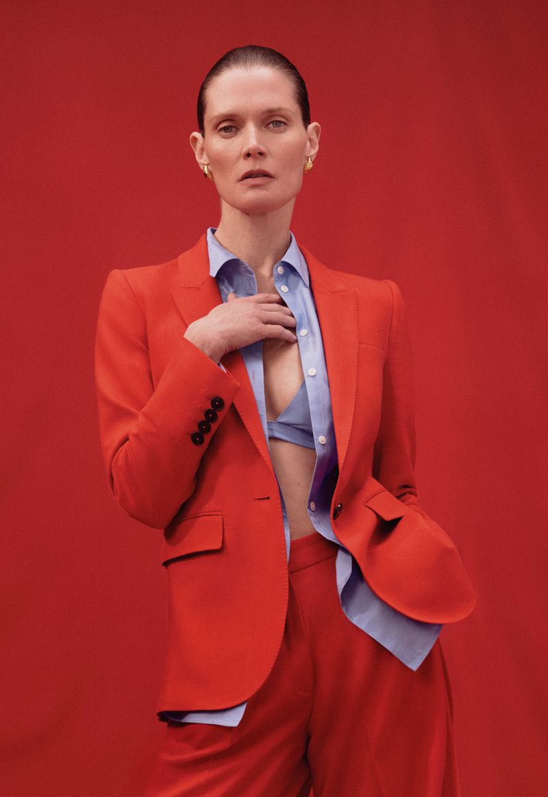 Fashionable woman with a red suit and blue shirt poses in a portrait against a matching red backdrop for a studio fashion shoot.
