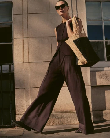 Malgosia Bela walking outdoors wearing a black sleeveless vest, matching wide-leg trousers, sunglasses, and a woven tote bag.