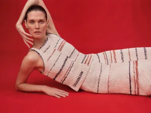 Model Malgosia Bela reclining on a bright red background wearing a cream knit sleeveless top and skirt