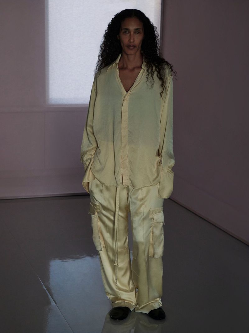 Model standing indoors wearing a light yellow silk button-down shirt and matching trousers in a softly lit room