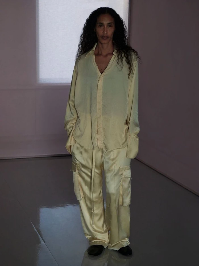 Model standing indoors wearing a light yellow silk button-down shirt and matching trousers in a softly lit room