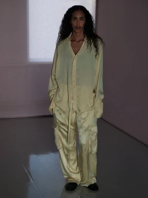 Model standing indoors wearing a light yellow silk button-down shirt and matching trousers in a softly lit room