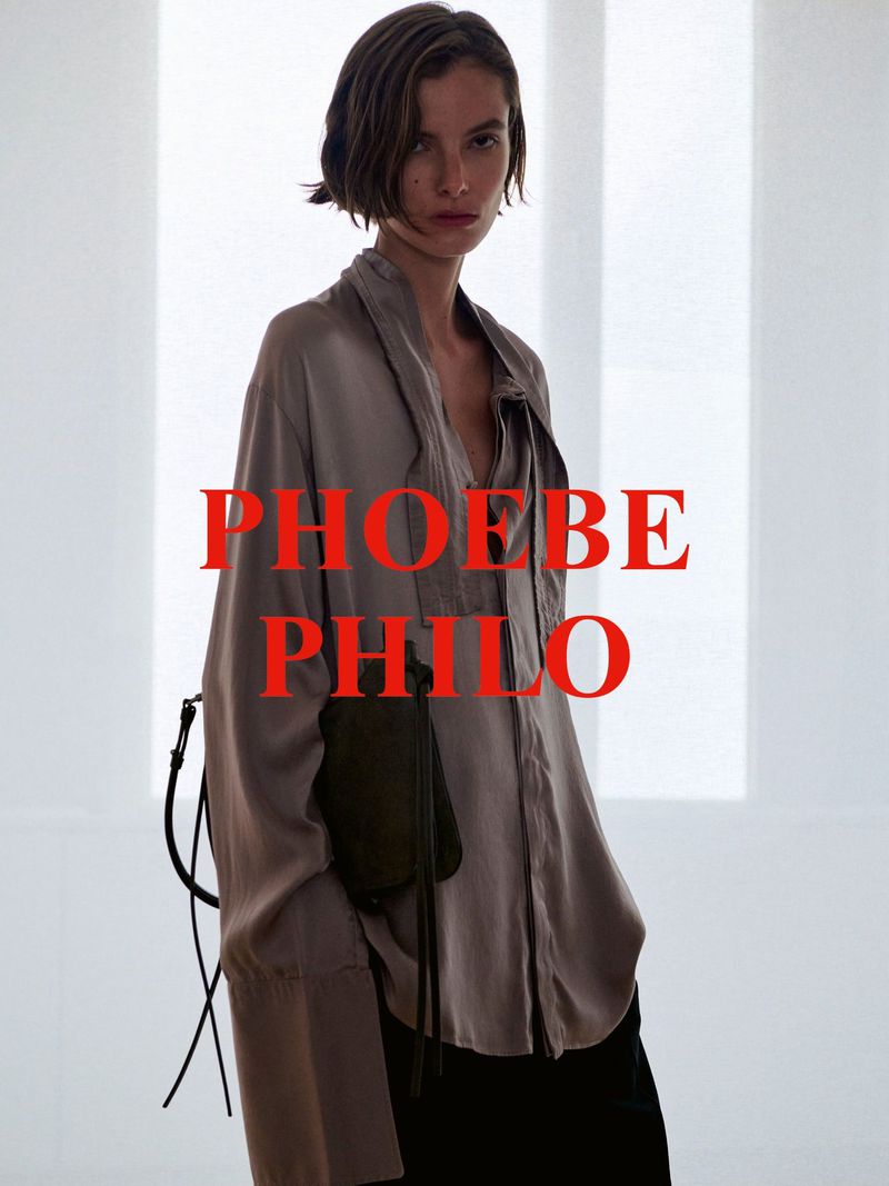 Model wearing a loose taupe silk shirt and carrying a dark shoulder bag against a bright minimalist studio background.