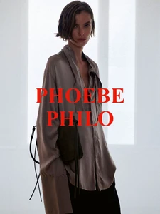 Model wearing a loose taupe silk shirt and carrying a dark shoulder bag against a bright minimalist studio background.