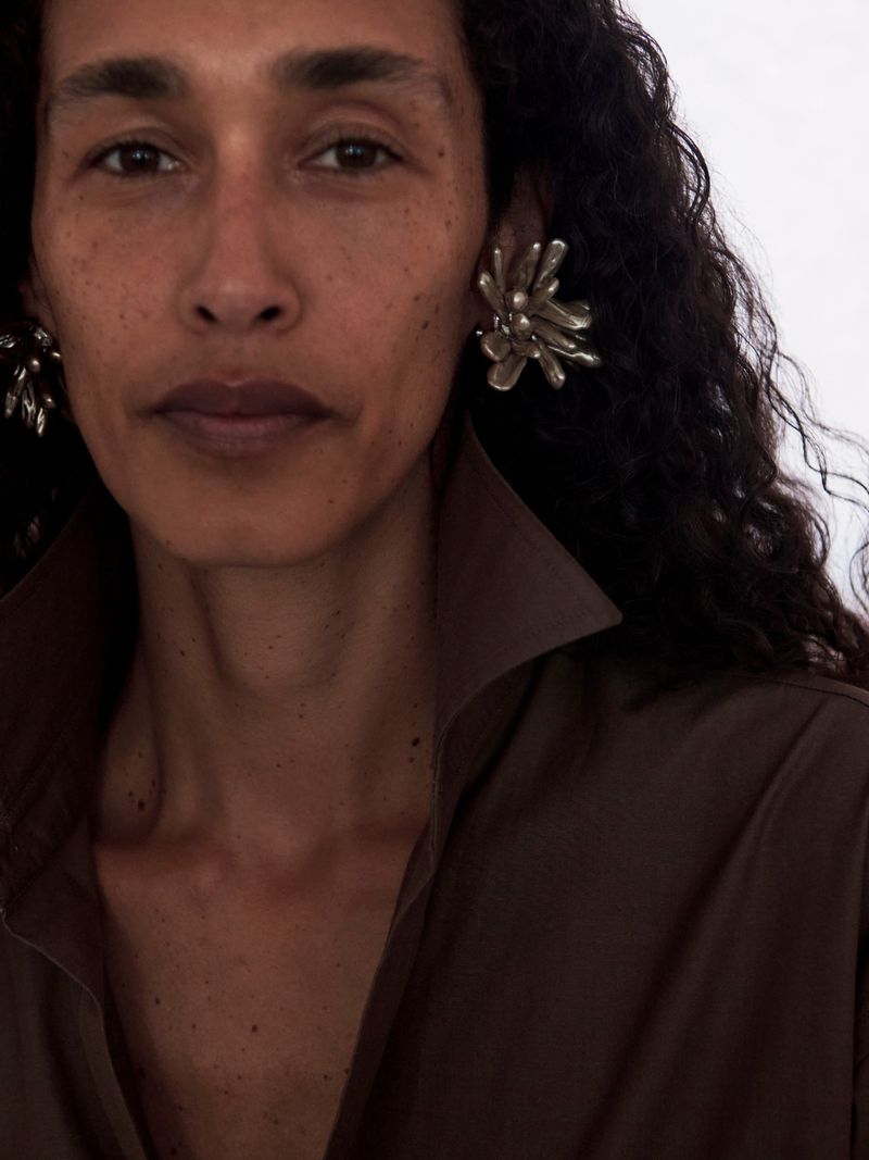 Close-up portrait of a woman with dark curly hair wearing sculptural silver earrings and a brown collared shirt against a white background.