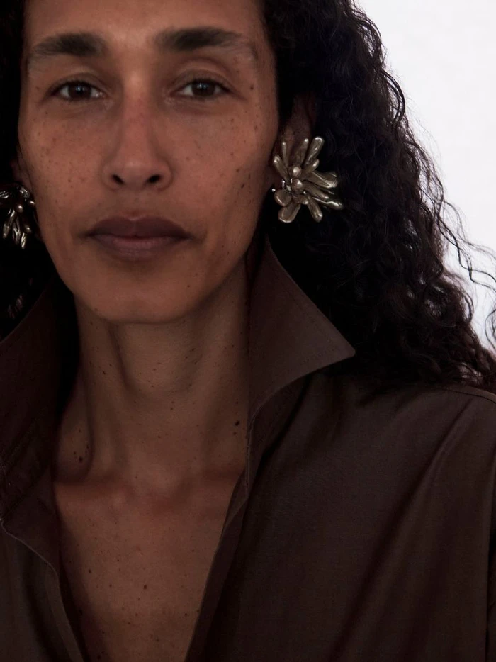 Close-up portrait of a woman with dark curly hair wearing sculptural silver earrings and a brown collared shirt against a white background.