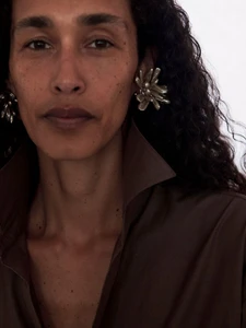 Close-up portrait of a woman with dark curly hair wearing sculptural silver earrings and a brown collared shirt against a white background.