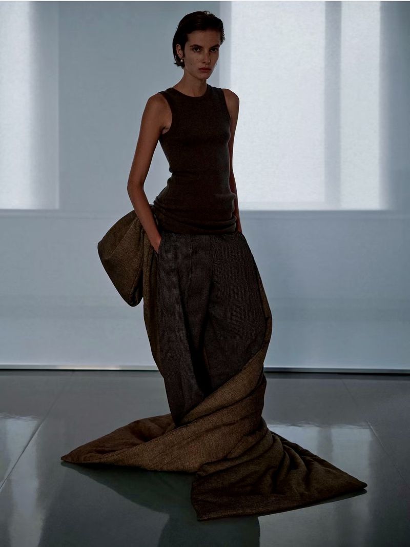 Model standing on a reflective floor wearing a dark tank top and trousers wrapped in a long brown fabric for Phoebe Philo Collection F.