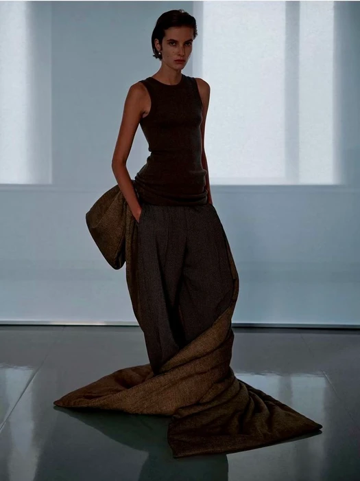 Model standing on a reflective floor wearing a dark tank top and trousers wrapped in a long brown fabric for Phoebe Philo Collection F.