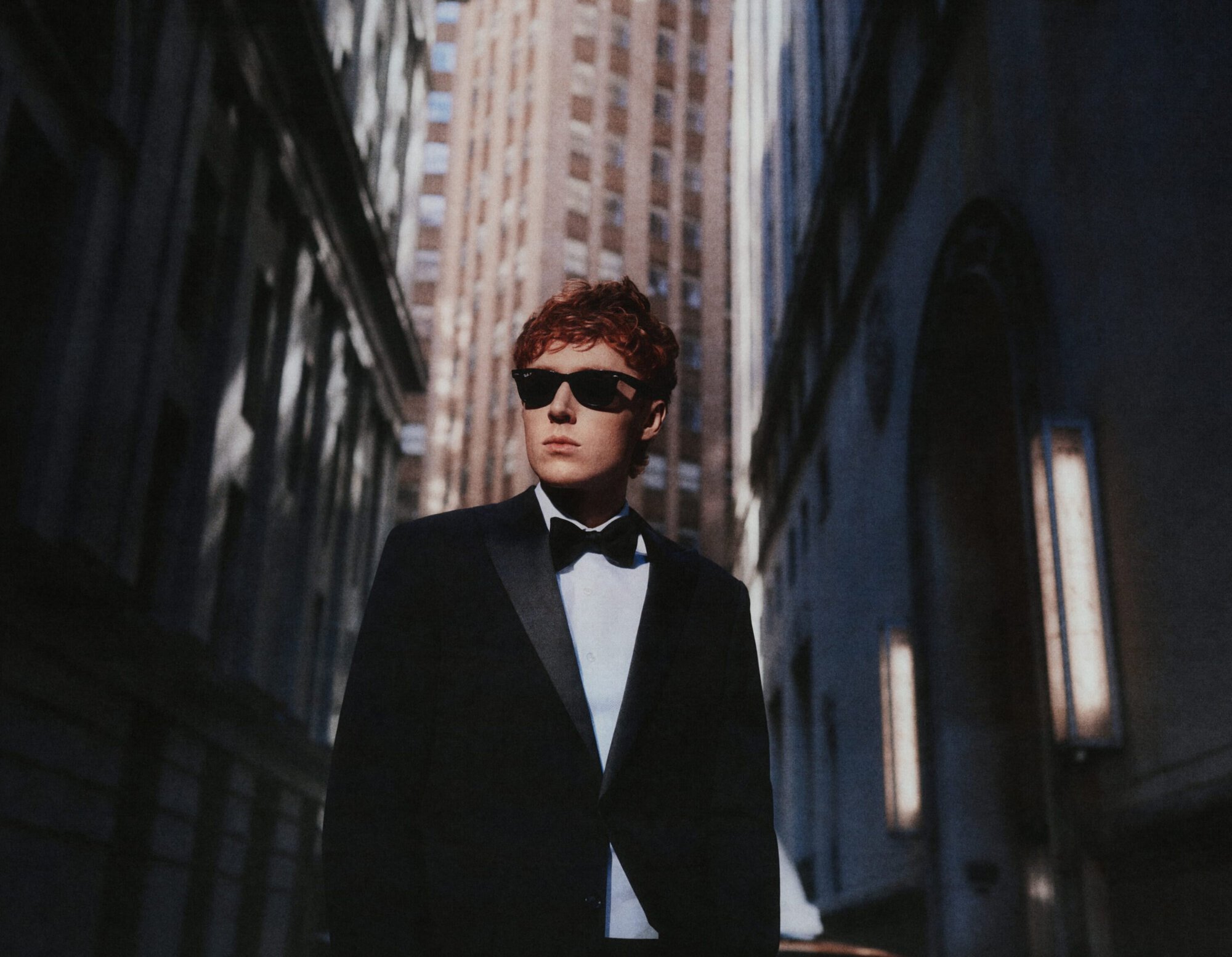 Man wearing a black tuxedo and sunglasses standing in a canyon between tall city buildings