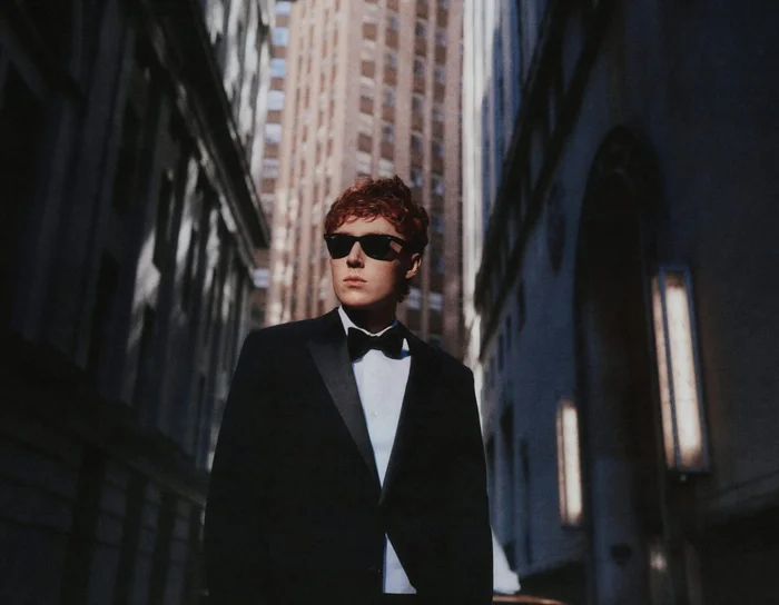Man wearing a black tuxedo and sunglasses standing in a canyon between tall city buildings