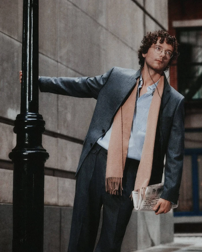 Man with curly hair in a grey suit and camel scarf leaning against a black lamppost on a city street