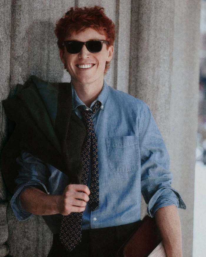 Smiling red-haired man wearing sunglasses, blue button-down shirt, patterned tie, and holding a jacket against a stone pillar