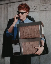 Man with red hair wearing sunglasses holding two cardboard boxes and talking on a phone against stone wall