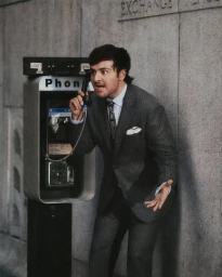 Man in a grey pinstripe suit shouting on a public payphone outdoors against a stone wall.