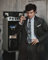 Man in dark pinstripe suit and paisley tie talking on public payphone against stone wall