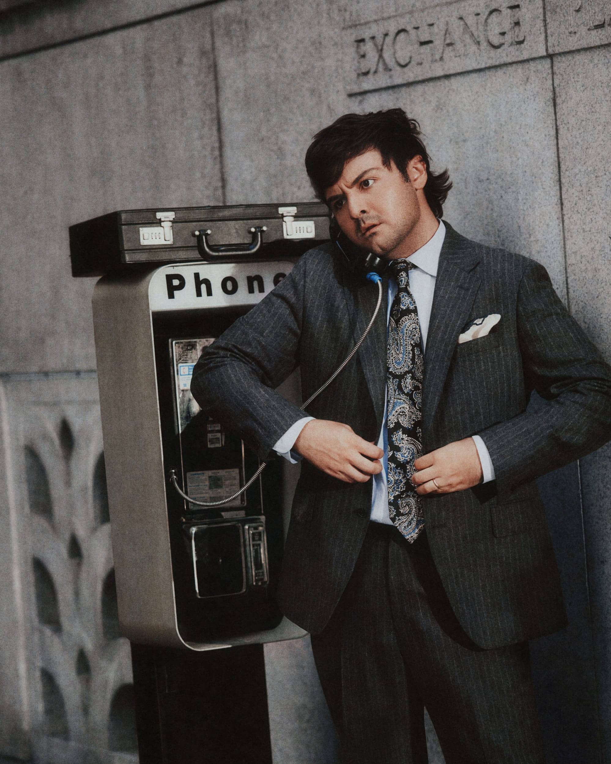 A man in a pinstripe suit talking on an outdoor payphone next to a stone wall.