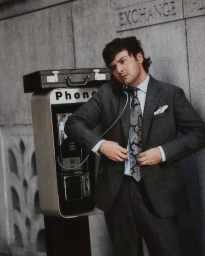 A man in a pinstripe suit talking on an outdoor payphone next to a stone wall.