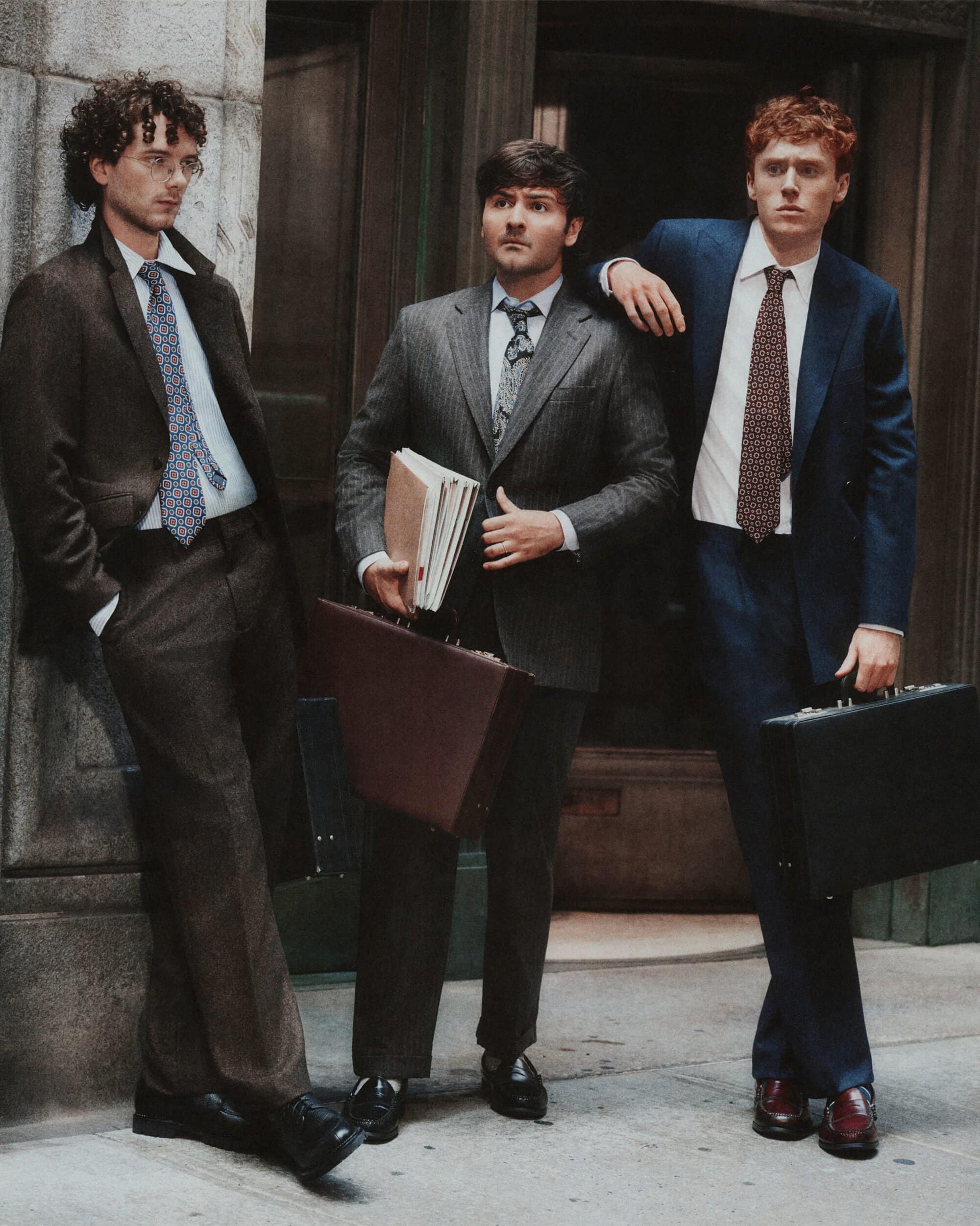 Three men in tailored suits holding briefcases and folders outside a building for the J. Crew Fall 2026 ad campaign.