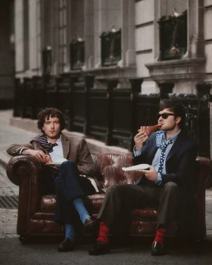 Two men in blazers seated on a leather sofa on a city street, one eating a slice of pizza