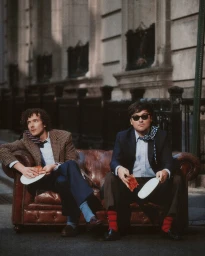 Two men seated on a leather chesterfield sofa outdoors holding plates with pizza slices for J. Crew Fall 2026 campaign.