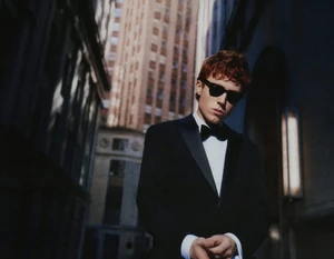 Man wearing a black tuxedo, bow tie, and sunglasses standing in a city street between tall buildings