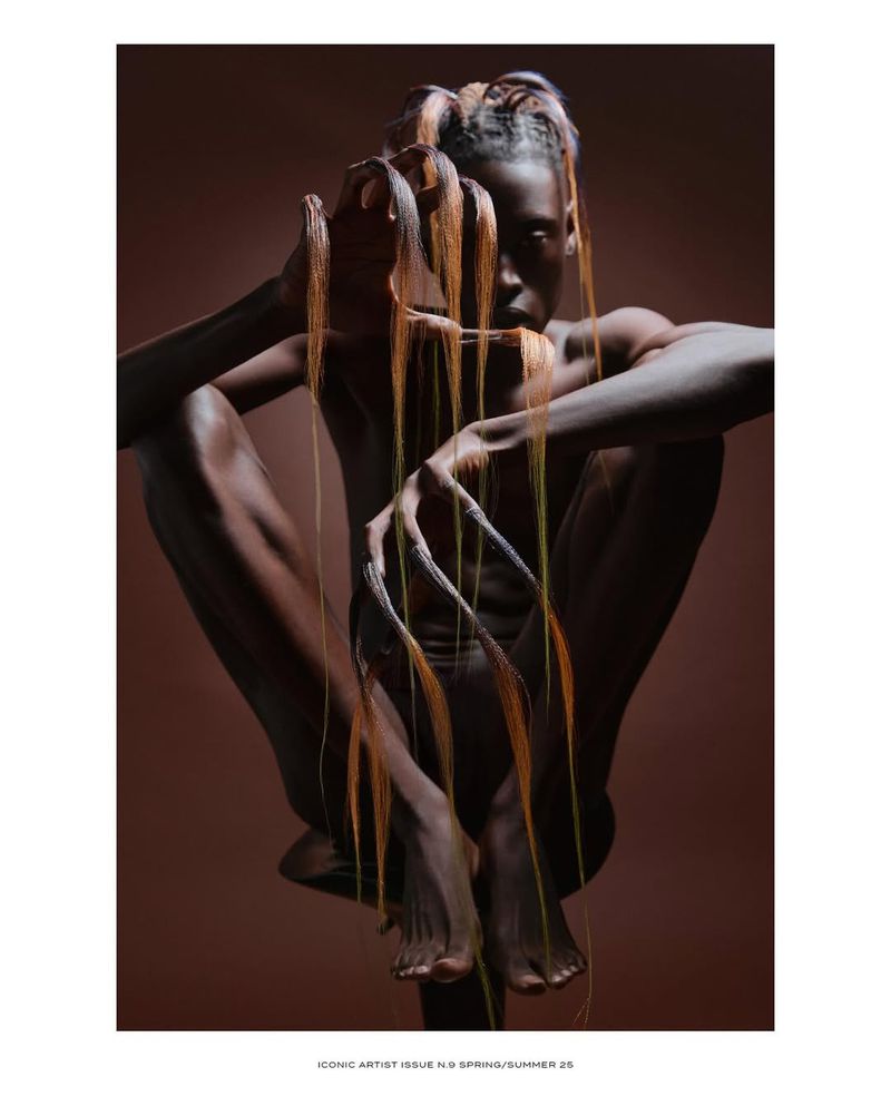 Striking studio portrait of an adult female with sculptural hair and nail extensions, posing in front of a solid brown backdrop.