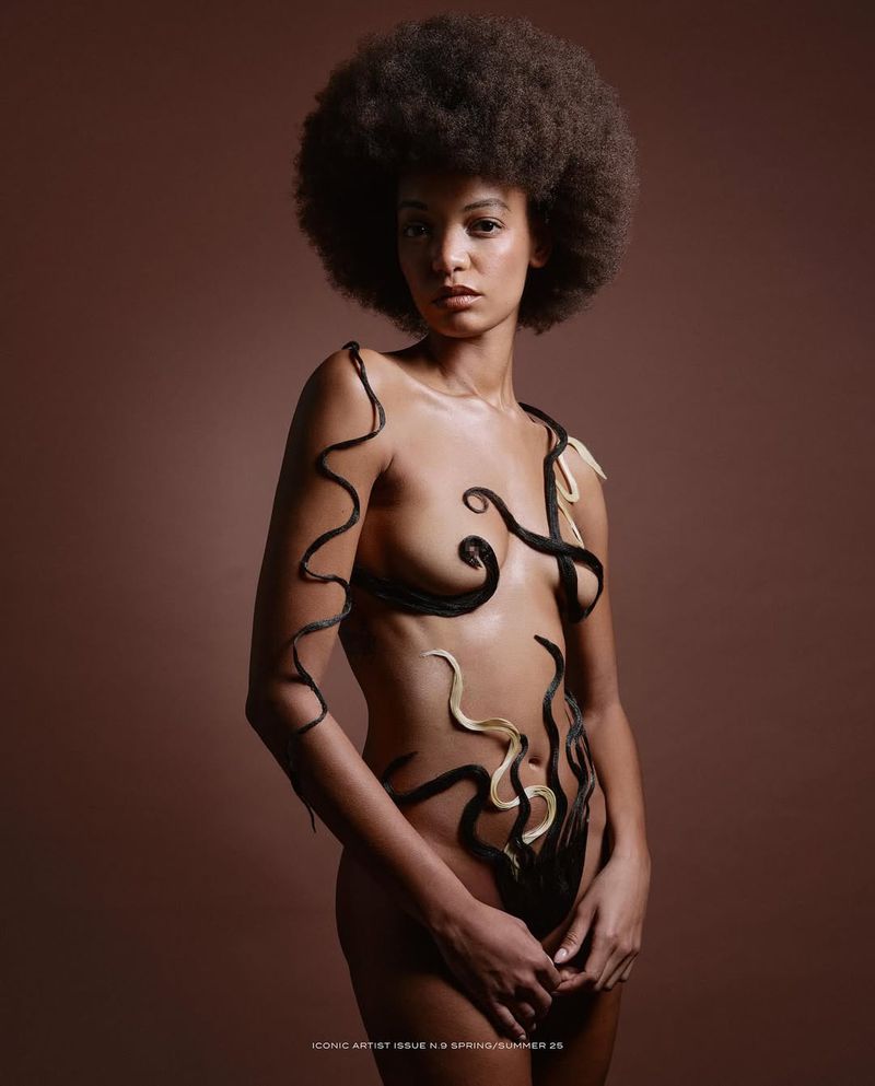 Artistic portrait of a Black woman with an afro, posing nude with hair strands forming creative designs on her body against a brown background.