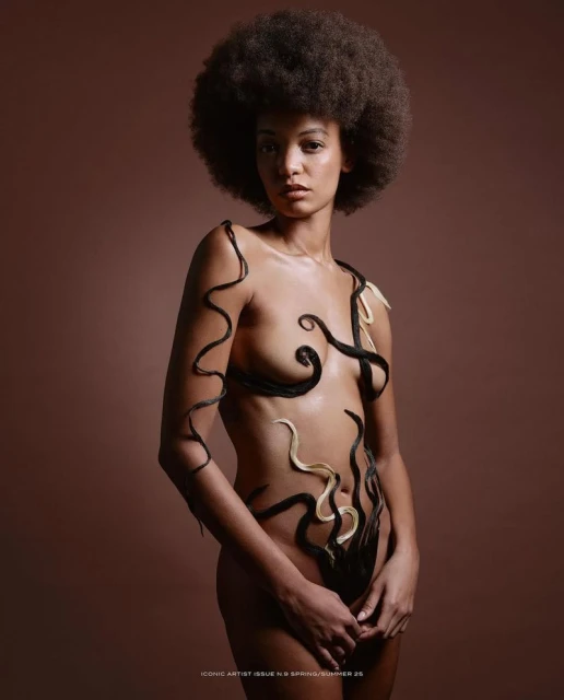 Artistic portrait of a Black woman with an afro, posing nude with hair strands forming creative designs on her body against a brown background.
