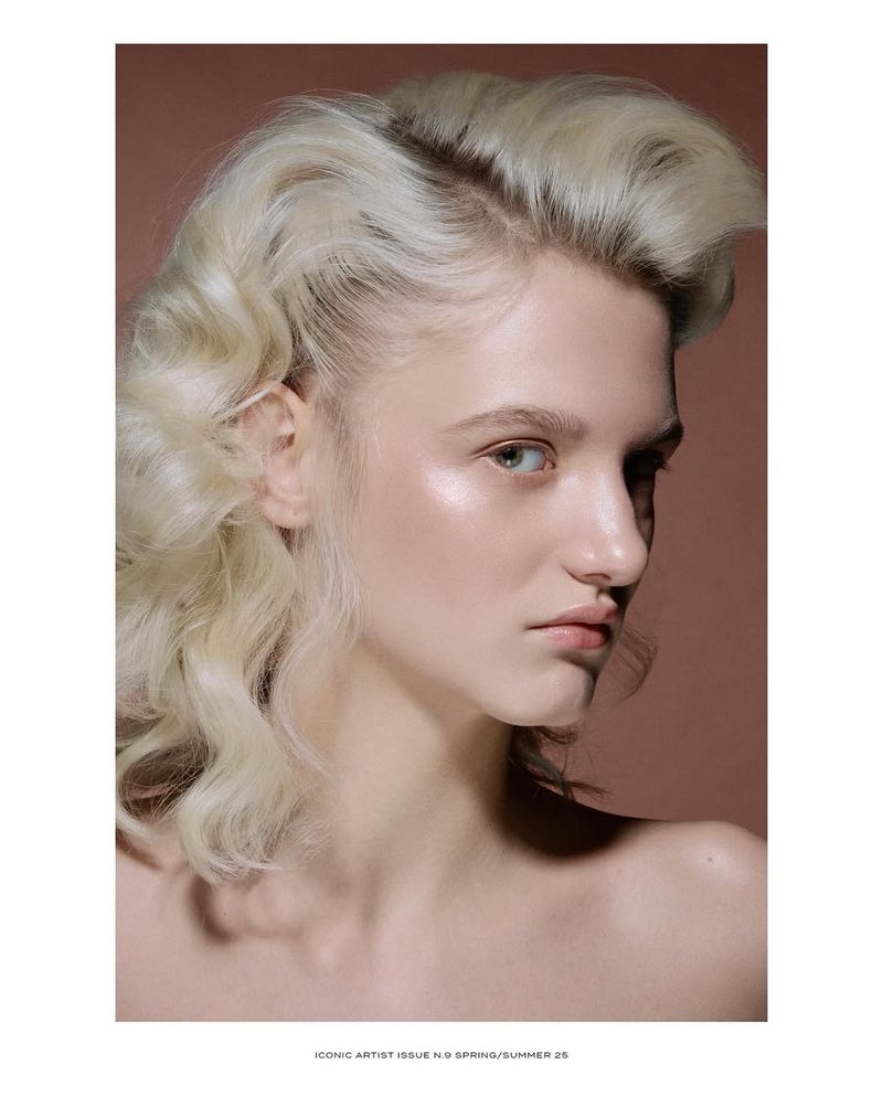Beauty shot of a pale-skinned woman with styled blonde hair, wearing minimal makeup against a brown background in portrait orientation.