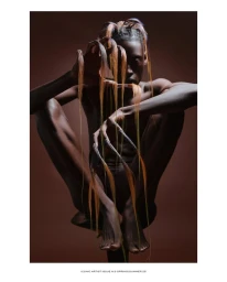Model crouching in a sculptural pose with long orange and brown hair strands extending from their hands against a brown backdrop.