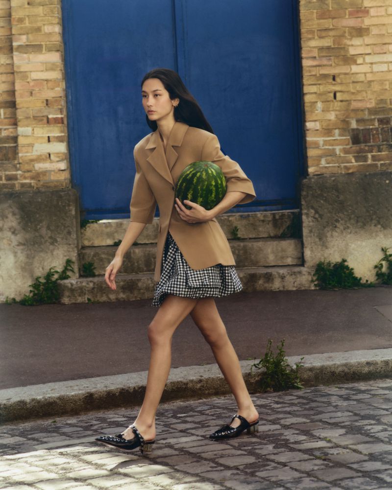 Woman walks on cobblestone street carrying watermelon in a tan blazer and black and white dress, with a blue door in the background.