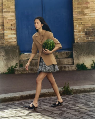 Woman walks on cobblestone street carrying watermelon in a tan blazer and black and white dress, with a blue door in the background.
