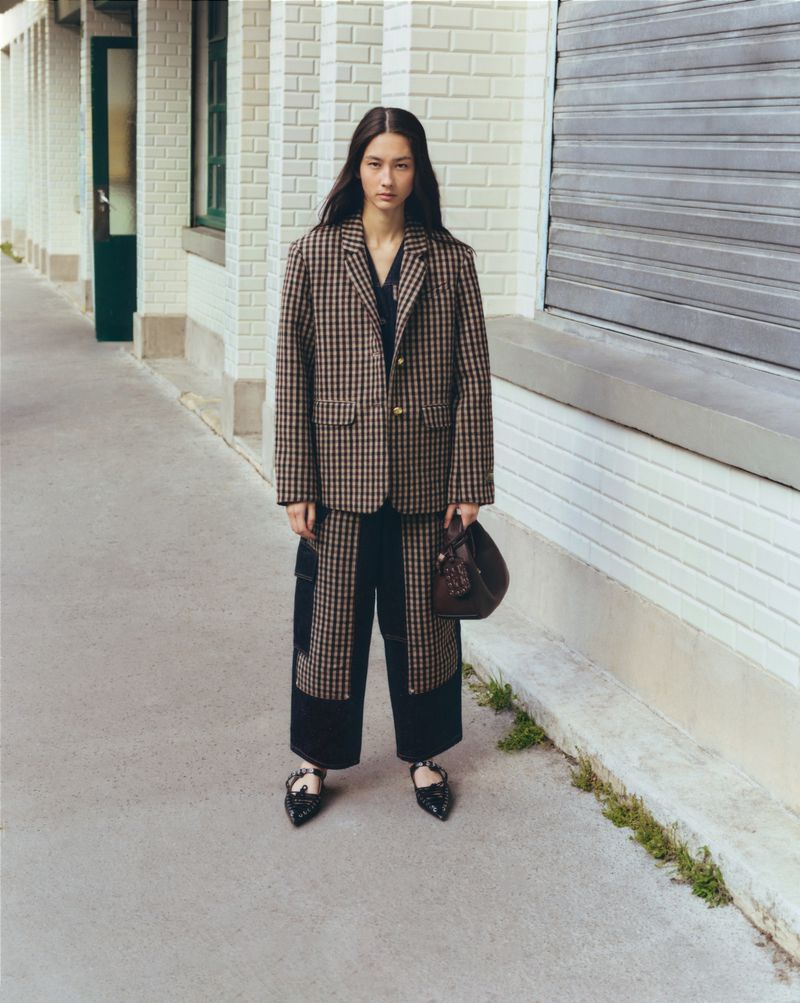 A stylish woman in a plaid blazer and dark pants stands outside on a concrete path next to a white brick building with a metal door.