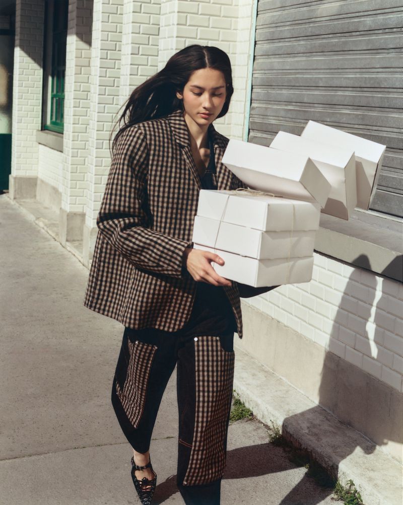 A woman in a checked blazer carries a stack of white boxes on a city street with a closed garage door and a brick building.