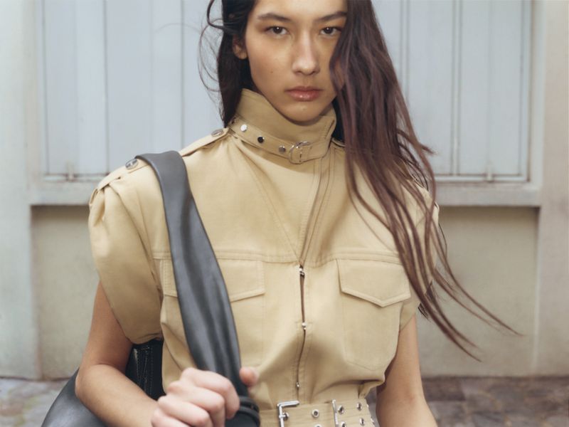 Portrait of a woman with long brown hair wearing a tan utility-style jacket and carrying a gray bag against a blurred light wall.