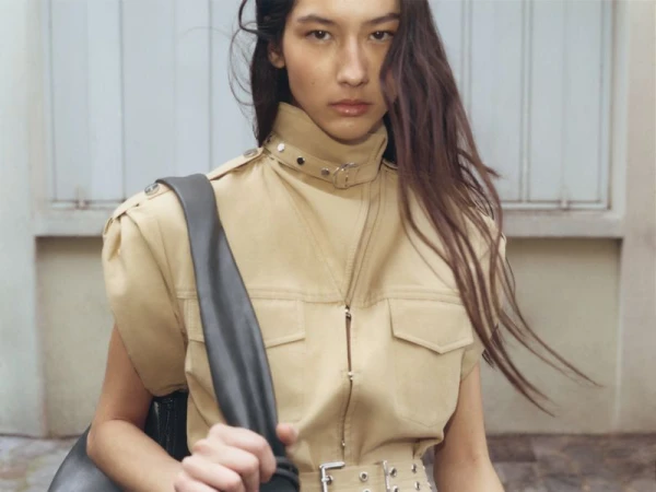 Portrait of a woman with long brown hair wearing a tan utility-style jacket and carrying a gray bag against a blurred light wall.