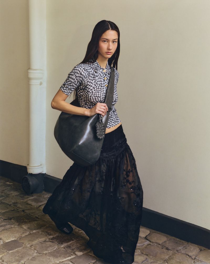 Fashionable woman poses in a patterned blouse, long sheer skirt, and black leather purse in a bright indoor setting.
