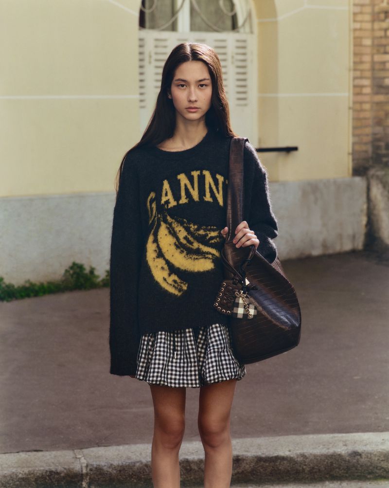 A fashionable young woman poses outdoors in a black GANNI sweater with bananas, a plaid skirt, and a brown purse against a building backdrop.
