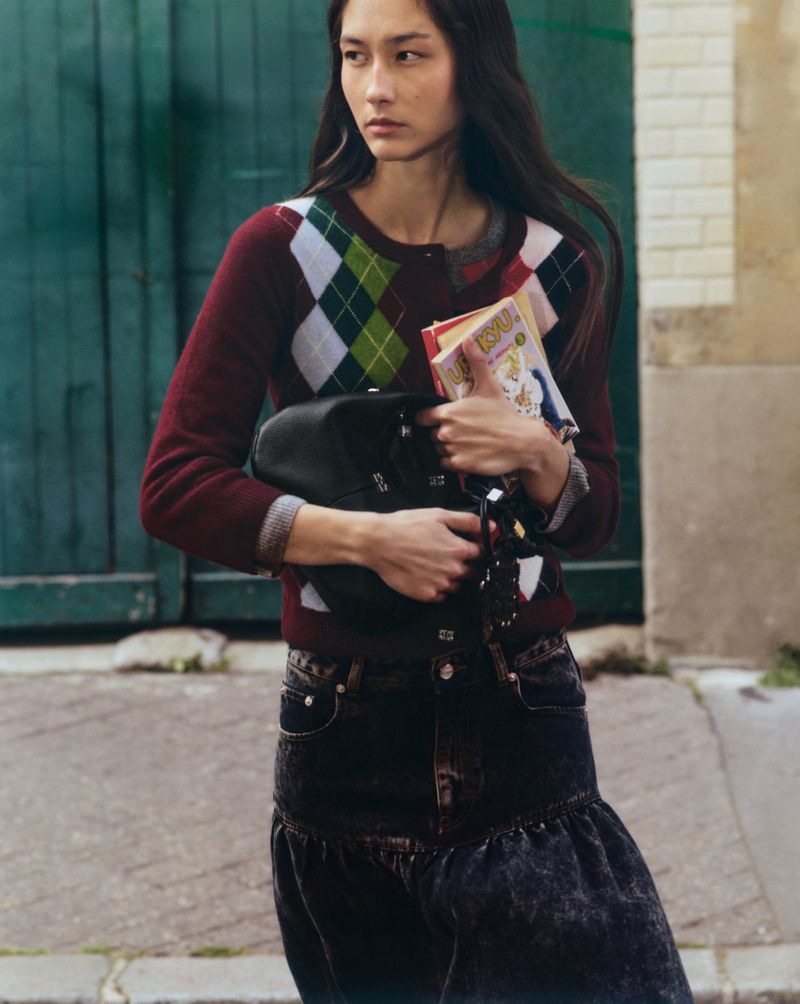 An Asian woman stands outdoors with a burgundy argyle sweater, denim skirt, and black bag, holding books in front of a green door.