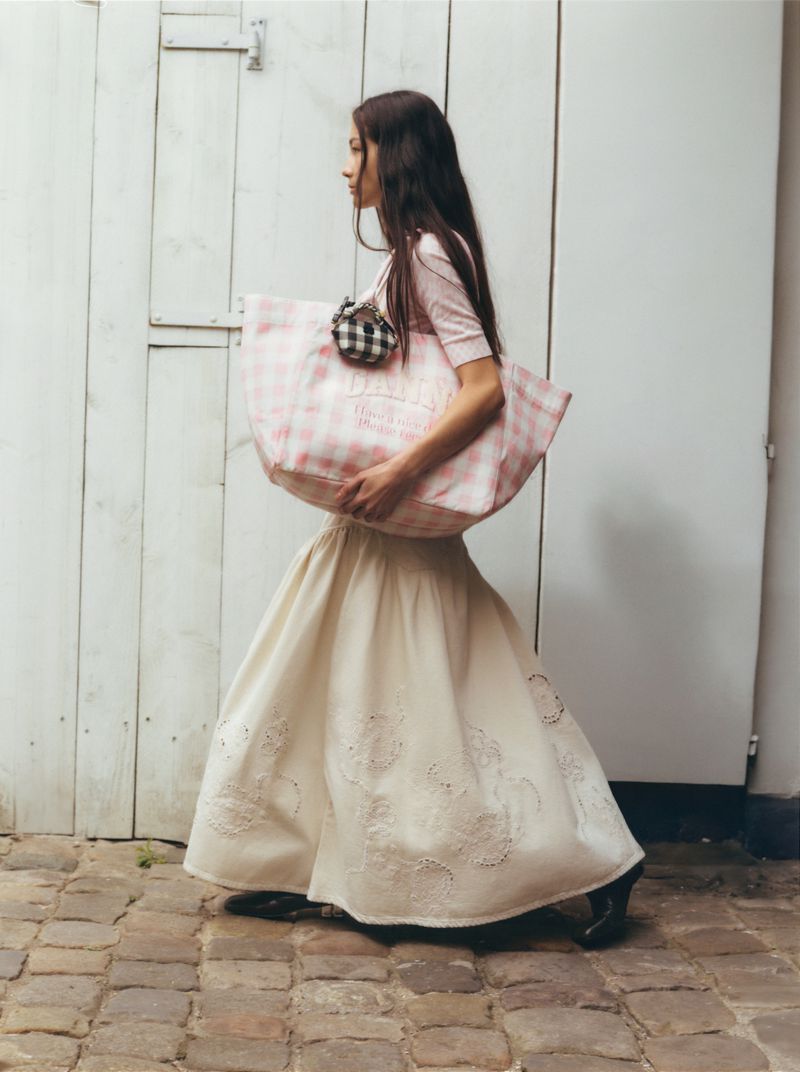 Woman in cream skirt and pink top carries pink gingham tote bag outdoors near white wooden door and brick pavement.